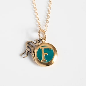 Small gold round locket, with initial F set in teal blue resin, with a brass flamingo charm. On a cable chain on a white background.