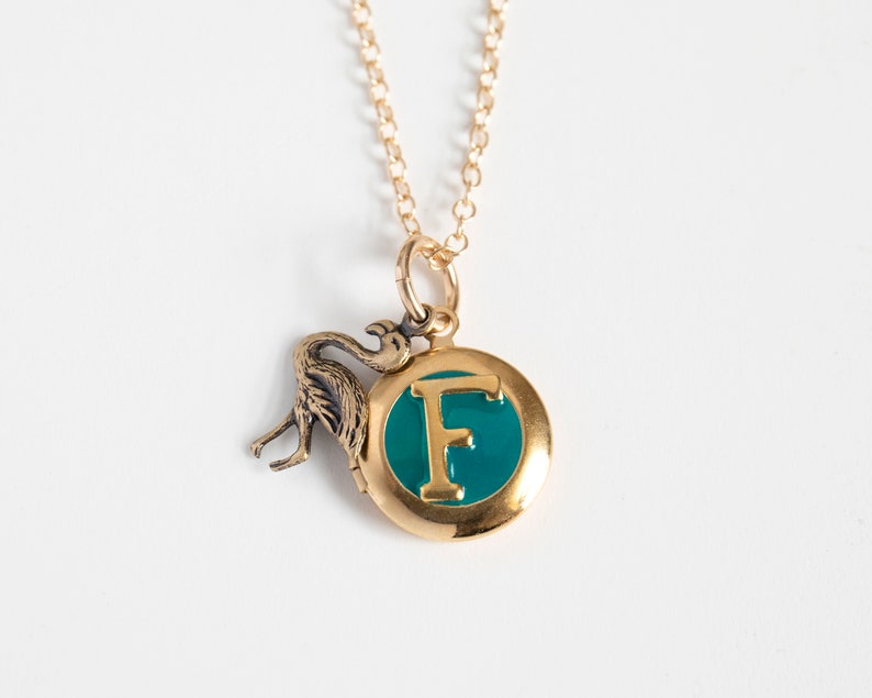 Small gold round locket, with initial F set in teal blue resin, with a brass flamingo charm. On a cable chain on a white background.