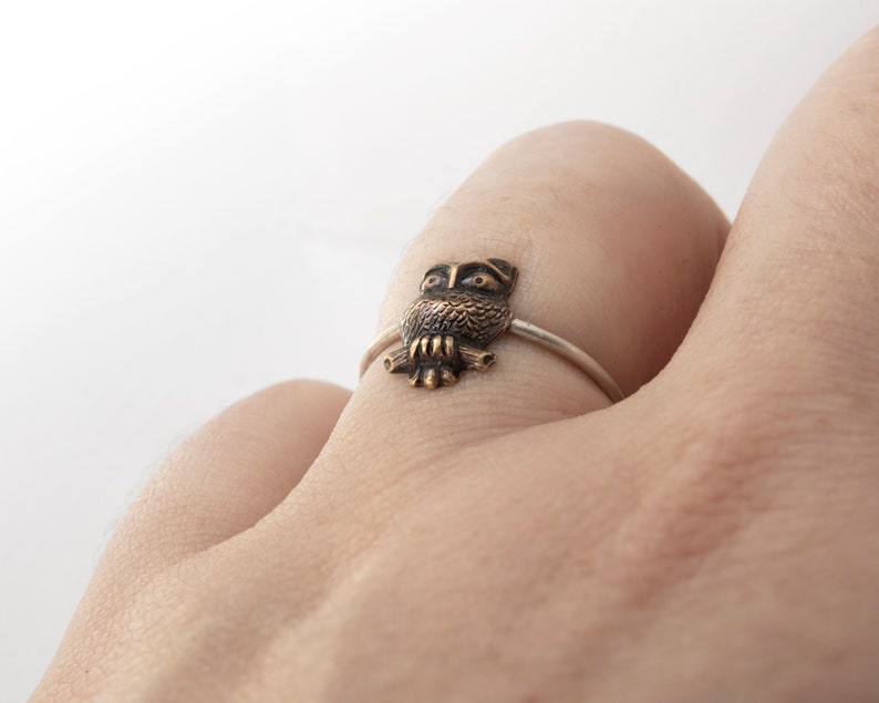 Tiny brass owl ring with a thin silver band. Worn on a hand on a white background.