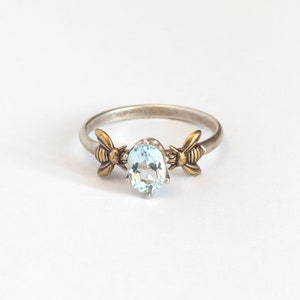Sky blue oval gemstone ring, in a prong setting with a brass bee on each side. Shown on a white background.