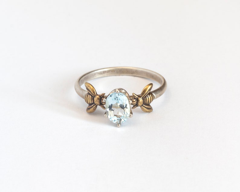 Sky blue oval gemstone ring, in a prong setting with a brass bee on each side. Shown on a white background.