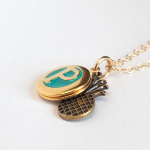 Small gold round locket, with initial P set in teal blue resin, with a brass pineapple charm. On a cable chain at an angle on a white background.