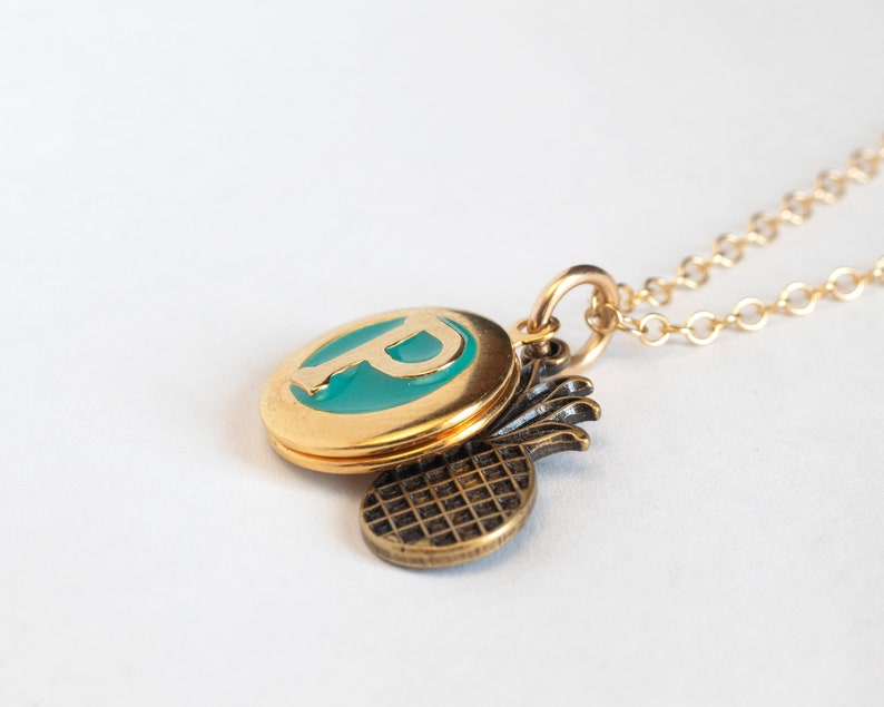 Small gold round locket, with initial P set in teal blue resin, with a brass pineapple charm. On a cable chain at an angle on a white background.