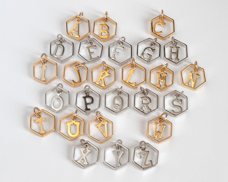 26 hexagon pendants with an initial inside, laid out in alphabetical order showing rows of silver or gold. on a white table