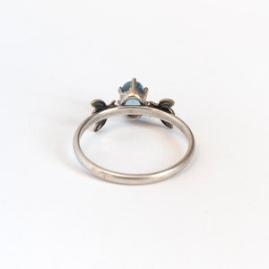 London blue oval gemstone ring, in a prong setting with a brass bee on each side. Shown from the back on a white background.