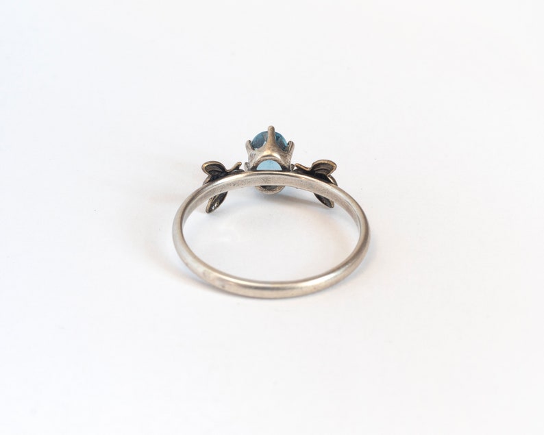 London blue oval gemstone ring, in a prong setting with a brass bee on each side. Shown from the back on a white background.