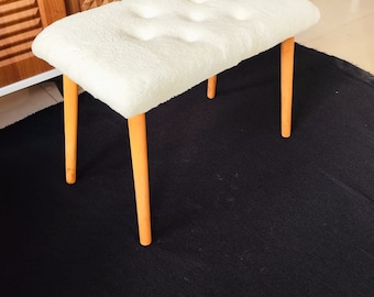 May include: A small, rectangular ottoman with a plush, off-white cushion and four orange-colored wooden legs. The ottoman is a decorative furniture piece, suitable for a living room or bedroom. The legs are approximately 15 inches tall.