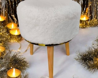 May include: A small, round, white faux fur ottoman with three wooden legs. The ottoman has a yellow and black patterned trim. The background includes candles and tinsel.