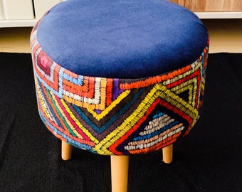 May include: A small, round stool with a navy blue velvet seat and a vibrant, embroidered skirt. The skirt displays a geometric design in red, yellow, green, and blue. It has three wooden legs. The stool's height is approximately 40 cm.