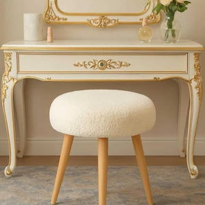 May include: A white vanity table with gold trim, a matching gold-framed mirror, and a round stool with a white textured fabric seat and three light wood legs. The vanity features a single drawer with a central gold floral ornament.