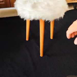 以下が含まれることがあります： ふわふわの白いトップと3本の木製の脚が付いた、小さくて丸いスツール。脚は明るいオレンジ色です。スツールは黒い表面にあります。