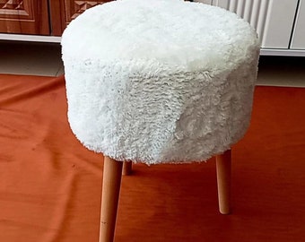 May include: A small, round white faux fur ottoman with three light brown wooden legs. The plush, fluffy top contrasts with the smooth, tapered legs. This decorative footstool is a cozy addition to any living space.