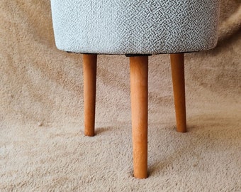 May include: A small, round ottoman with a light gray and white patterned fabric. It has three wooden legs and a small decorative item on top. The ottoman is on a beige shag rug.