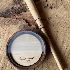 May include: A wooden coaster and a matching wand-shaped stirrer. The coaster has a dark brown rim, a clear center, and a light wood interior. The stirrer is light-colored wood with dark accents. The coaster is inscribed with "Heartwood 2024".