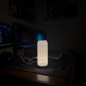 May include: A white Monster Energy Ultra can illuminated as a lamp on a desk. The can is lit from within, displaying the Monster Energy logo and text. The desk also has a keyboard, mousepad, and computer monitors in the background.