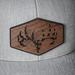May include: A gray baseball cap with a hexagonal wooden patch featuring a black and white deer antler design.