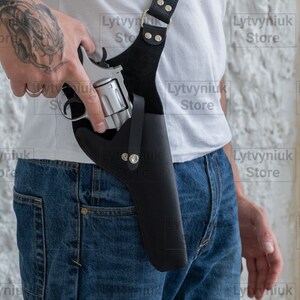 Può includere: Una fondina a spalla in pelle nera contenente un revolver in metallo color argento. La fondina è dotata di cinghie regolabili, rivetti metallici e una chiusura a bottone automatico, progettata per tenere l'arma verticalmente contro il busto.
