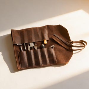 Pode incluir: Um estojo de ferramentas em couro castanho escuro com vários bolsos interiores e um fecho de atar. O estojo contém várias ferramentas manuais de metal, incluindo brocas, uma pequena chave de fendas e um punção, organizados em ranhuras.