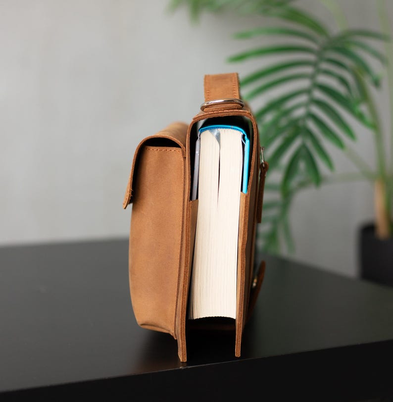 May include: A brown leather satchel bag with a book inside. The bag has a flap closure and a metal buckle. The bag is sitting on a black surface.