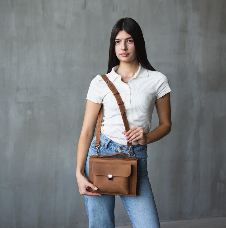 May include: A woman wearing a white collared shirt and blue jeans is carrying a brown leather satchel bag with a long strap.