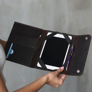 May include: A brown leather tablet case with a built-in pen holder and multiple pockets. The case is open and a tablet is visible inside.