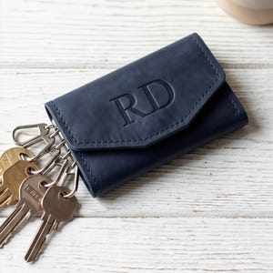May include: A navy blue leather key holder with a flap closure and the embossed letters RD on the front. The key holder features multiple metal key hooks attached to the interior, with several brass-colored keys hanging from the side of the case.