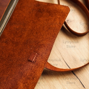 K&ouml;nnte beinhalten: Nahaufnahme einer braunen Ledertasche mit einem Futter aus Wildleder. Die Tasche hat einen Klappverschluss und einen Riemen. Der Riemen ist aus dem gleichen Leder wie die Tasche. Die Tasche liegt auf einer Holzoberfl&auml;che.