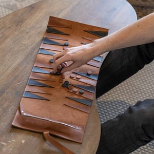 May include: A brown leather backgammon board with black and blue triangular accents. The board is rolled out on a wooden table, with a hand placing game pieces. The board has a zipper closure and a carrying strap.