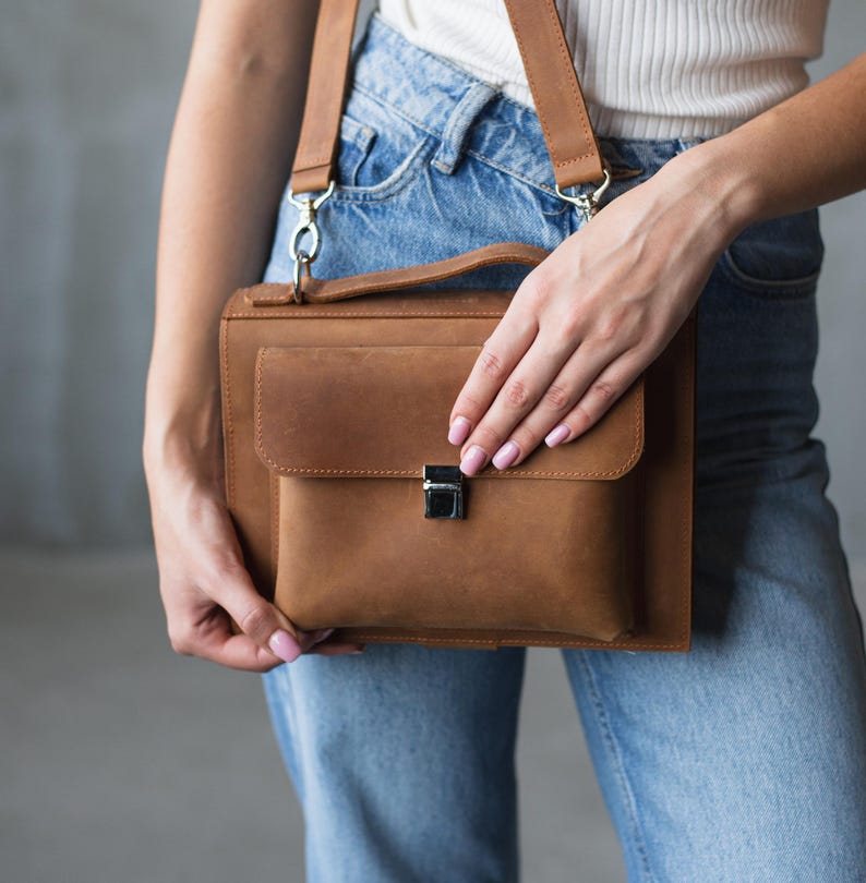 May include: A brown leather satchel bag with a silver clasp and a long strap. The bag is being held by a person wearing blue jeans.