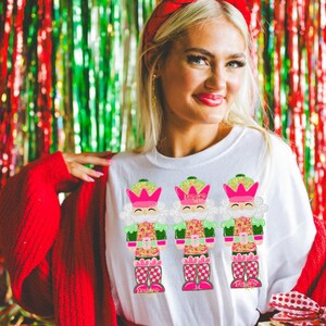 May include: White t-shirt with three vibrant nutcracker designs. The nutcrackers have pink crowns, green details, and patterned clothing. The background is a festive mix of red and green tinsel.