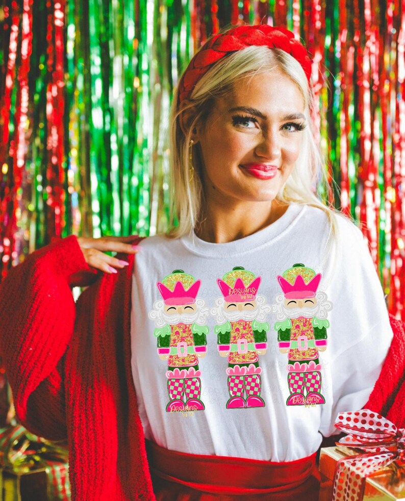 May include: White t-shirt with three vibrant nutcracker designs. The nutcrackers have pink crowns, green details, and patterned clothing. The background is a festive mix of red and green tinsel.