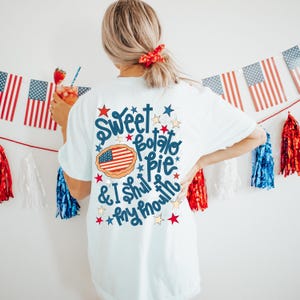 May include: White t-shirt with a blue and red graphic that says "Sweet Potato Pie & I Shut My Mouth". The graphic includes a pie with an American flag inside and stars.