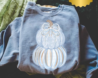 Puede incluir: Una sudadera azul de cuello redondo con un gráfico de dos calabazas apiladas. La calabaza superior es azul claro con un patrón de encaje blanco y un lazo decorativo, mientras que la inferior tiene rayas verticales azul claro y beige.