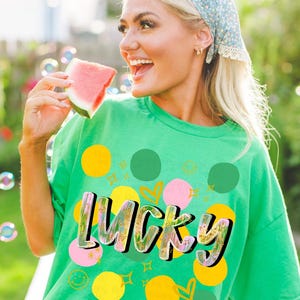 May include: A green t-shirt with the word "LUCKY" in a colorful, outlined font. The shirt features yellow, pink, and green polka dots, hearts, and smiley faces. The person is holding a slice of watermelon.
