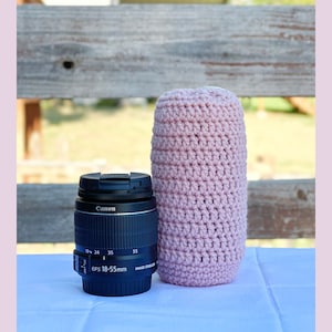 May include: A pink crocheted camera lens cozy with a Canon EF-S 18-55mm lens in front of it.
