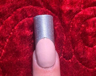 Puede incluir: Primer plano de un dedo con una punta de esmalte de uñas plateado. La uña está pintada de un color rosa claro y la punta tiene un acabado metálico plateado.