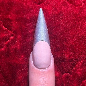 May include: A close-up of a finger with a long, pointed, silver nail with a nude base.