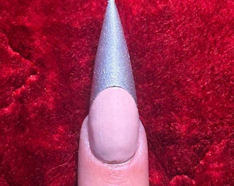 Puede incluir: Primer plano de un dedo con una uña larga, puntiaguda, plateada con una base nude.