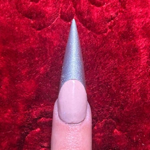 May include: A close-up of a finger with a long, pointed, silver nail with a nude base.