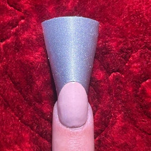 May include: A silver, cone-shaped nail form is placed over a painted fingernail. The nail is painted a light pink color. The background is a deep red, plush fabric.