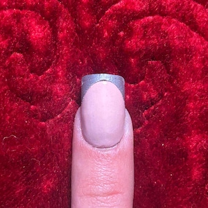 May include: A close-up of a fingernail with a light pink base and a silver metallic tip. The nail is on a finger, set against a red velvet background.