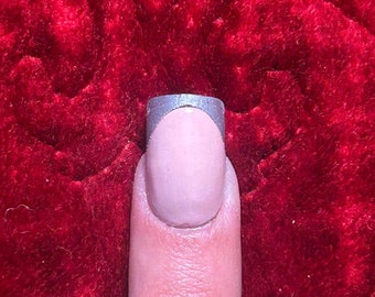 May include: A close-up of a fingernail with a light pink base and a silver metallic tip. The nail is on a finger, set against a red velvet background.