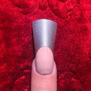 May include: A silver nail form is placed on a finger with a nude-colored nail. The nail form is cylindrical and has a slightly flared opening. The background is a textured, red surface.