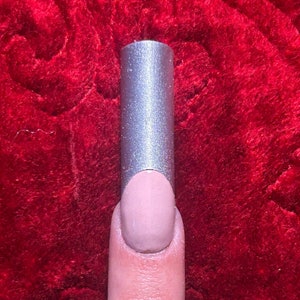 May include: A silver nail form is placed over a painted nail. The nail is painted a light pink color. The background is a red, textured fabric.