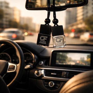 May include: Two black car air fresheners hanging from a rearview mirror. Each rectangular bottle has a black cap and the text "CF CLOUD FRAGRANCES". One bottle is black, the other is clear. The interior of a car is visible, with a blurred cityscape in the background.