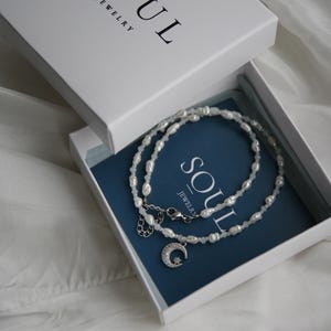 May include: A white box with the text "SOUL JEWELRY" on the top. The box is open and inside is a white pearl necklace with a silver moon and star charm. The necklace is on a blue background.