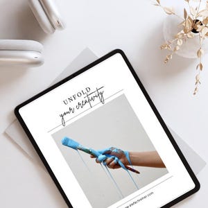 May include: A white tablet with a black border displays a graphic of a hand holding a paintbrush dripping blue paint. The text "UNFOLD your creativity" is displayed above the graphic. The website address "www.artoffactreiner.com" is displayed below the graphic.