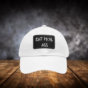Può includere: Cappellino da baseball bianco con una toppa rettangolare nera con la scritta bianca "EAT MOR ASS". Il cappellino è realizzato in un materiale morbido e ha una visiera curva. Lo sfondo è un muro scuro e strutturato.