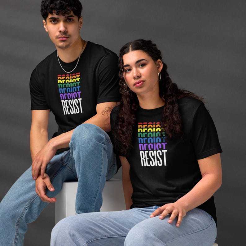 the word "RESIST" repeated in a cascading gradient of Pride Flag colors—red, orange, yellow, green, blue, and purple—culminating in a bold white "RESIST" at the bottom on a black unisex t-shirt worn by seated male and female models.