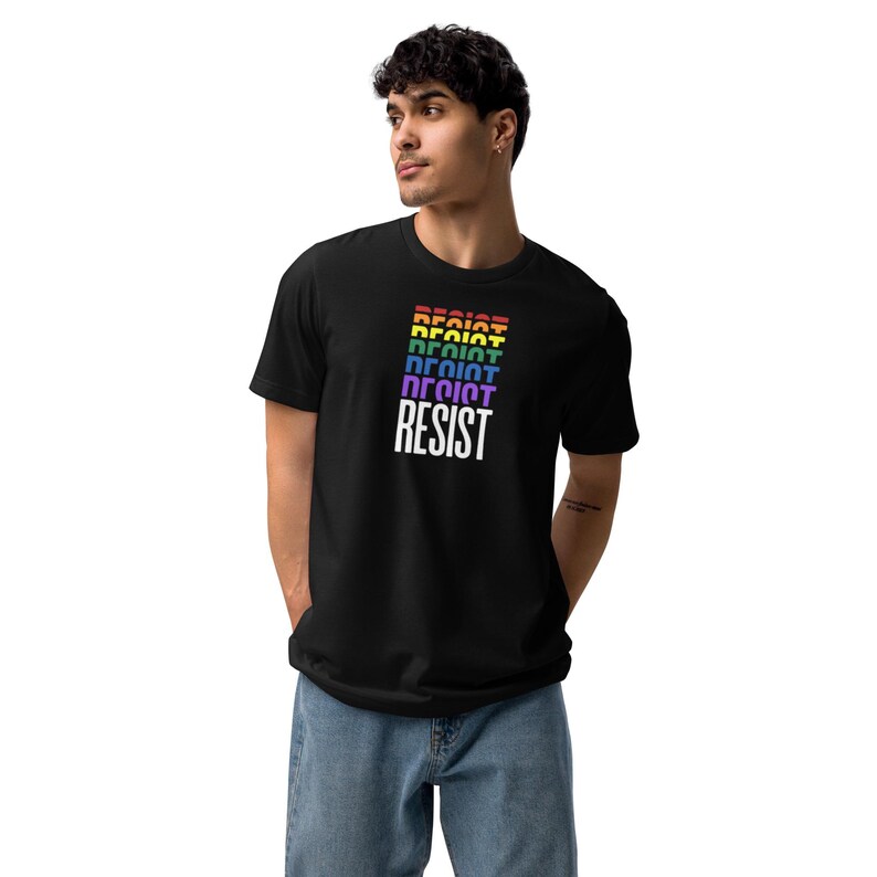 the word "RESIST" repeated in a cascading gradient of Pride Flag colors—red, orange, yellow, green, blue, and purple—culminating in a bold white "RESIST" at the bottom on a black unisex t-shirt worn by a standing male model